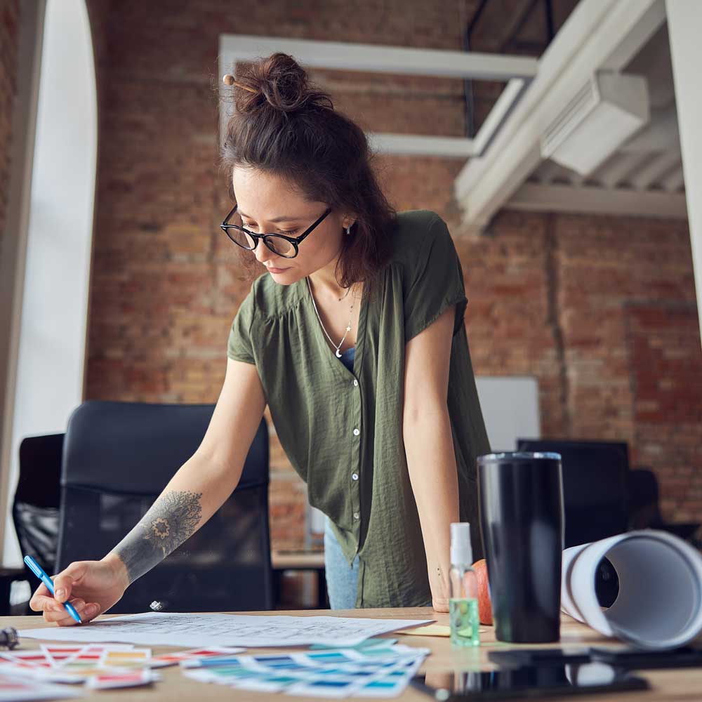 Creative woman, interior designer or architect wearing glasses making notes with a pen while working on a blueprint for new project, standing in her office. Home decoration and renovation, business